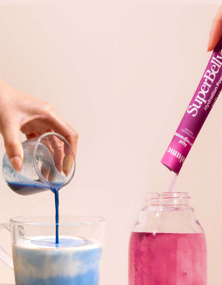 A person pours blue liquid from a small glass jug into a clear mug of frothy milk, creating a layered effect. Another hand dips a pink Super Belly hydration powder sachet into a mason jar filled with pink liquid.